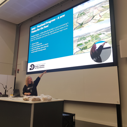 Rebekah stands behind a desk in the lecture hall at ARU pointing up at a large screen presenting Peatland Progress information