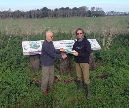 Andy and Henry stand in front of the interpretation boards at New Decoy, Henry hands Andy some books and they shake hands