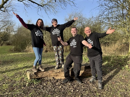 Two women stand on a log bench, two men stand in front, all arms outstretched smiling and wearing t-shirts that say Eat, Sleep, Restore, Re-Peat