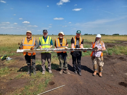 Five men in high vis waistcoats hold a long soil core, they stand in a flat fenland field, blue sky 