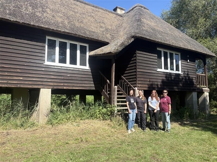 Four woman in front of a brown bungalow which is on wide stone stilts off the floor. The bungalow is made out of dark brown wood and it has a thatched roof