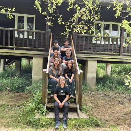 Multiple people sitting on some narrow steps that lead up to a bungalow. The dark brown bungalow is on high stone stilts.