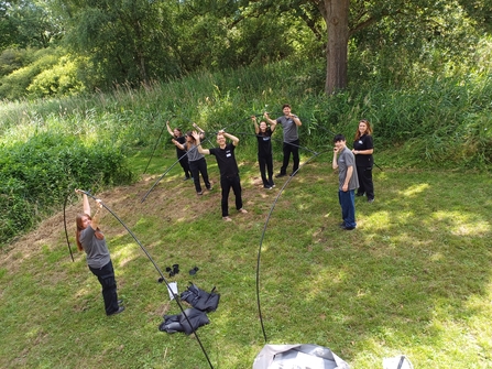 Multiple people holding up curved poles trying to assemble a giant tent. They're all looking up and smiling at a camera