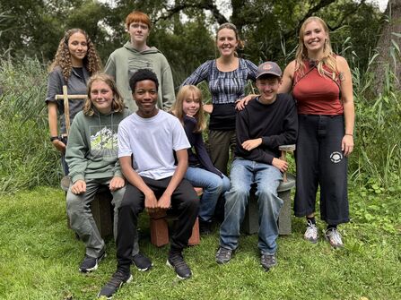 Five young people and three adults sit on and stand around handmade stools in a nature reserve