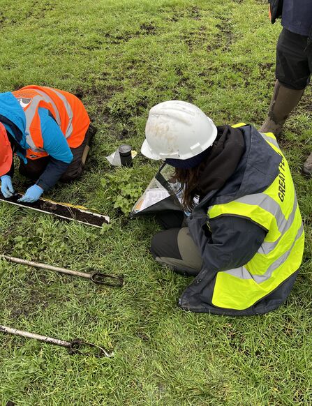 A young woman in a hard hat and high vis bent down on the ground filling out a form on a clipboard looking at a soil sample