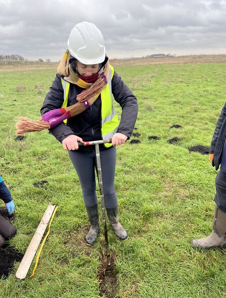 A young woman wrapped up in lots of layers and a scarf, wearing a hard hat and high vis holding an auger taking a sample of the soil