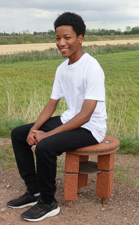 A teen Black boy sits on a red fibreboard stool with fields in the background