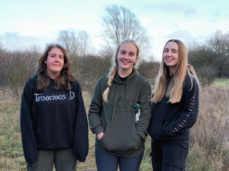 Kaitlyn, Katie and Ellen stand in a nature reserve, all wearing dark hoodies. Kaitlyn and Katie rest protective goggles on their heads