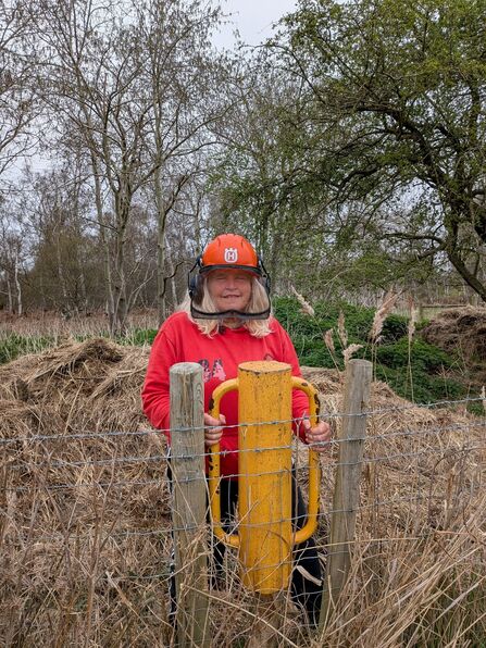 Catherine wears a helmet and red jumper using a fence post driver
