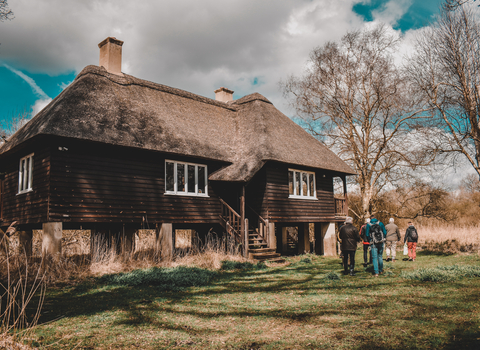 Rothschild's wooden bungalow on concrete stilts at Woodwalton Fen, with green grass in foreground and  blue sky and white clouds above. People look at it.