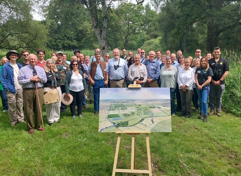 Group of 40 adults standing in a group smiling to camera, in front of them is a landscape watercolour on a wooden easel