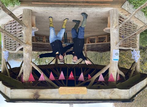 Two ladies stand with arms open wide in the wooden outdoor shelter