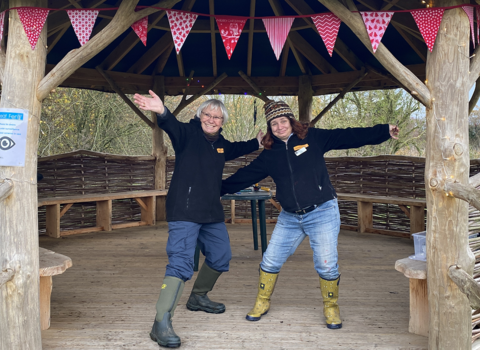 Caroline and Claire, wearing blue jeans, black great fen branded fleeces and wellies, stand arms outstretched inside a wooden octagonal shelter that's decorated with red bunting.